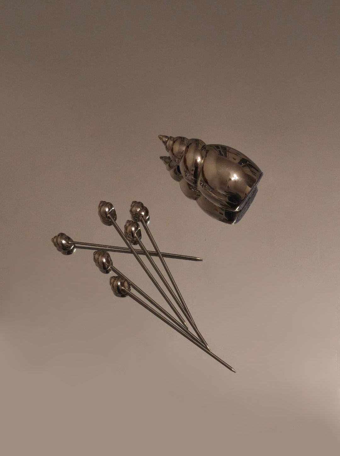Les Objoies Shell Cocktail Picks feature a silver, conch-shaped shell holder beside six ocean-inspired picks with round decorative tips, displayed on a plain light brown surface.