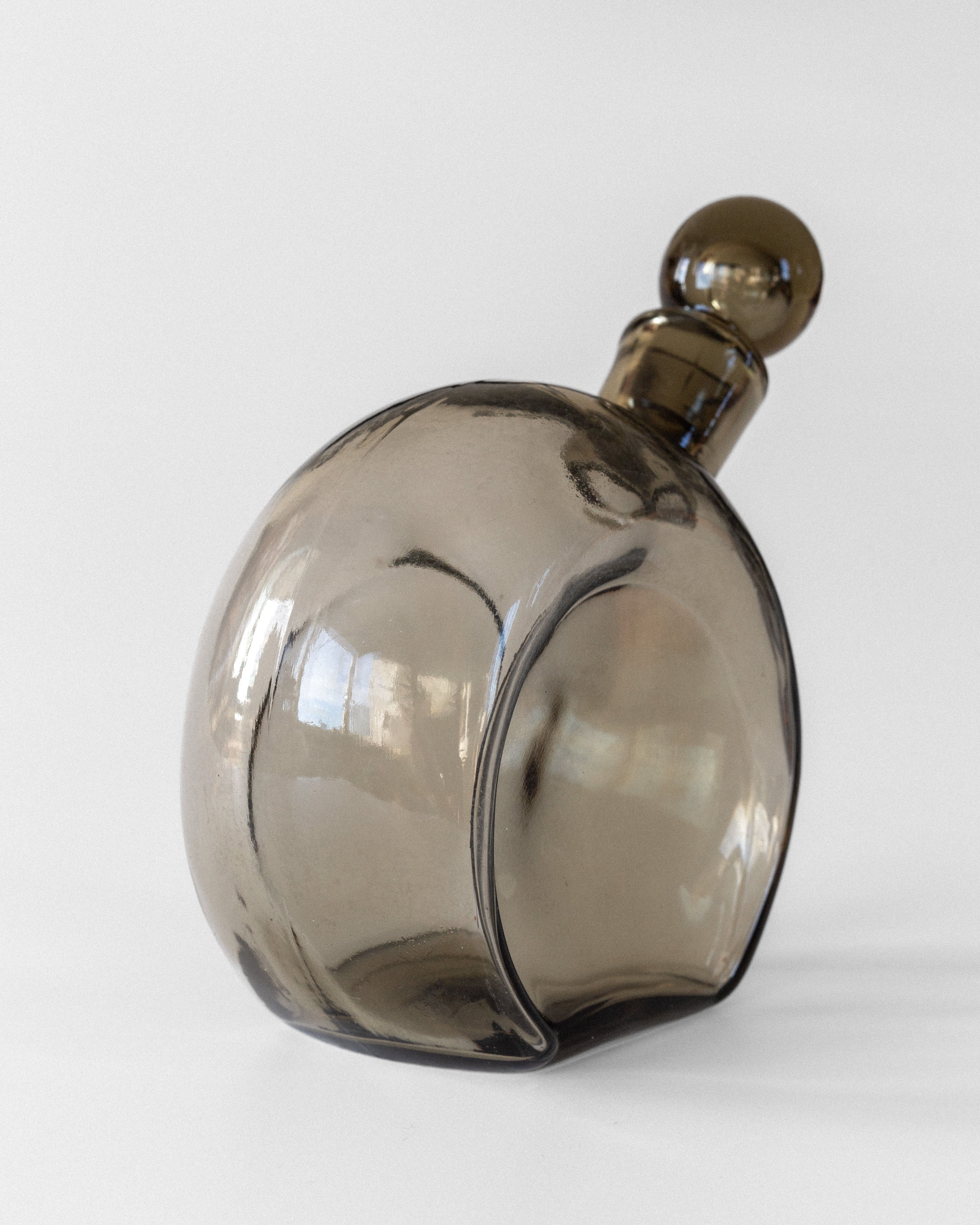 The Glass Decanter by Harald Notini, 1930s from Spigel showcases Swedish modernism with its transparent, curvy design and smooth smoky brown glass. Resting on its side on a white surface, the round stopper complements the decanter's reflective look and light-reflecting elegance.