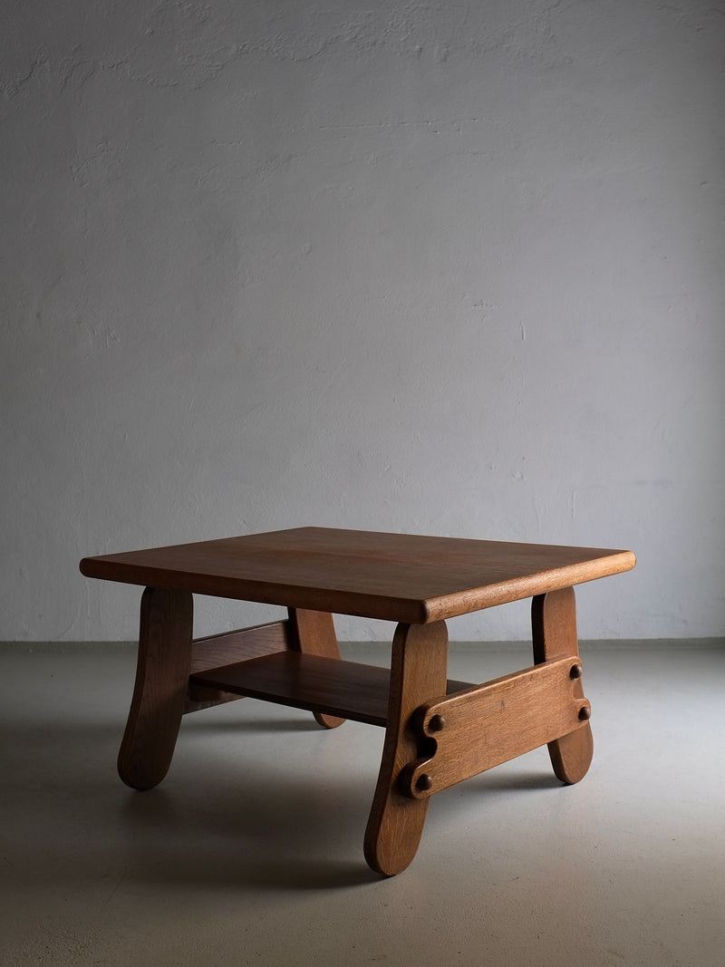 A small Brutalist Oak Coffee Table | Netherlands | 1970s with a simple, 1970s design stands against a plain, light gray wall. The table has a square top, curved legs, and a lower shelf. The lighting casts soft shadows, highlighting the table's natural wood grain and craftsmanship. This piece is from Veter Vintage.