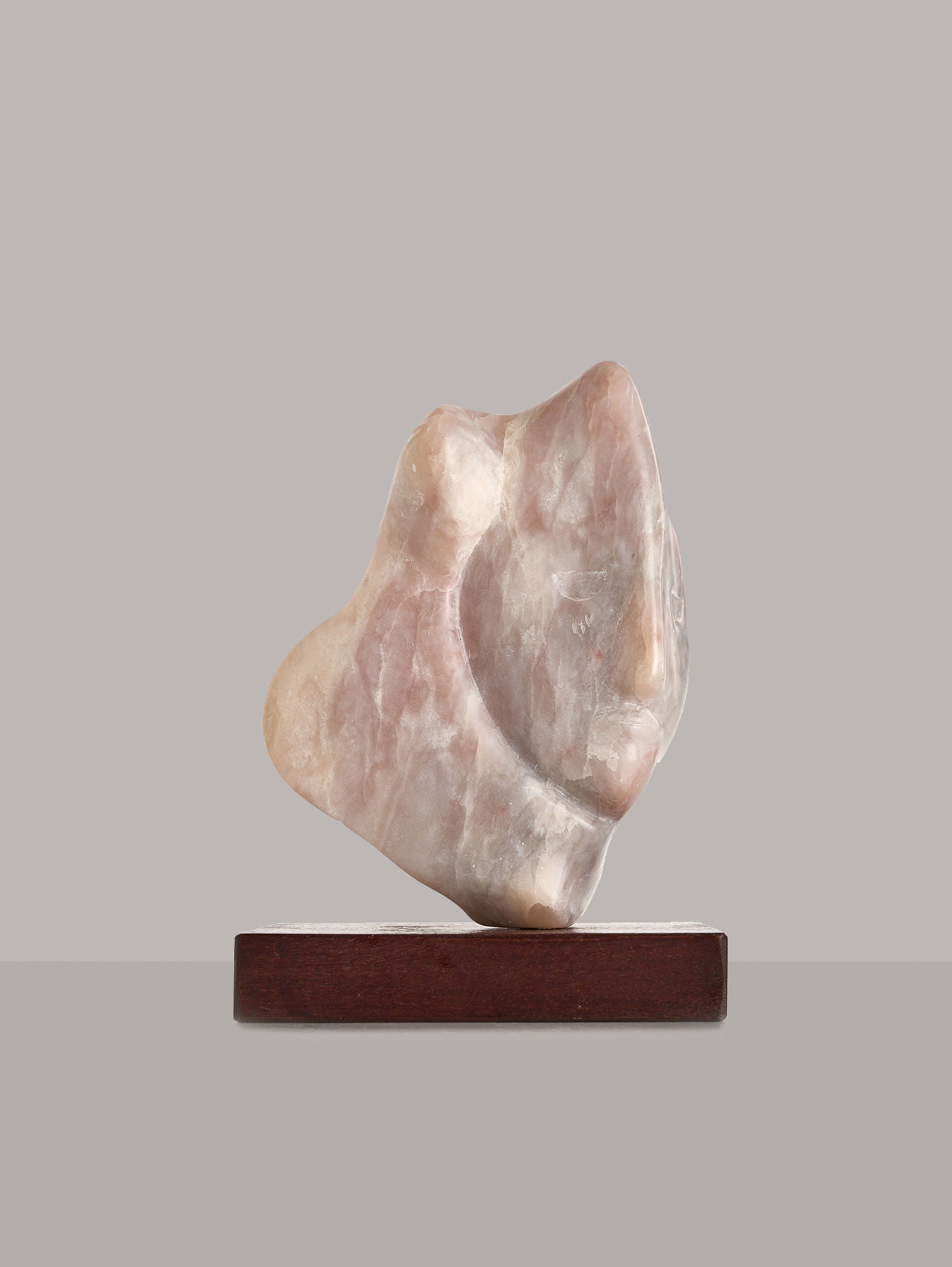 The Sculpture by Gisela Peterhof-Heimig, offered by RELIC LONDON, features an abstract marble design reminiscent of a female head with smooth curves and a blend of beige and light pink hues, elegantly positioned on a wooden plinth against a plain gray background.