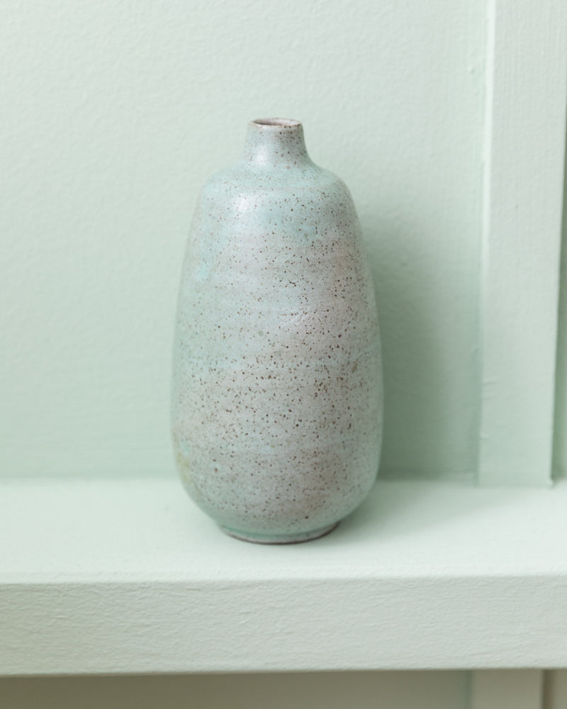 The Mint-Colored Vase by Carl Fischer (Bottega Jacobs, 1960s) with a slender neck and speckled finish sits on a white shelf against a pale wall, showcasing classic mid-century style.
