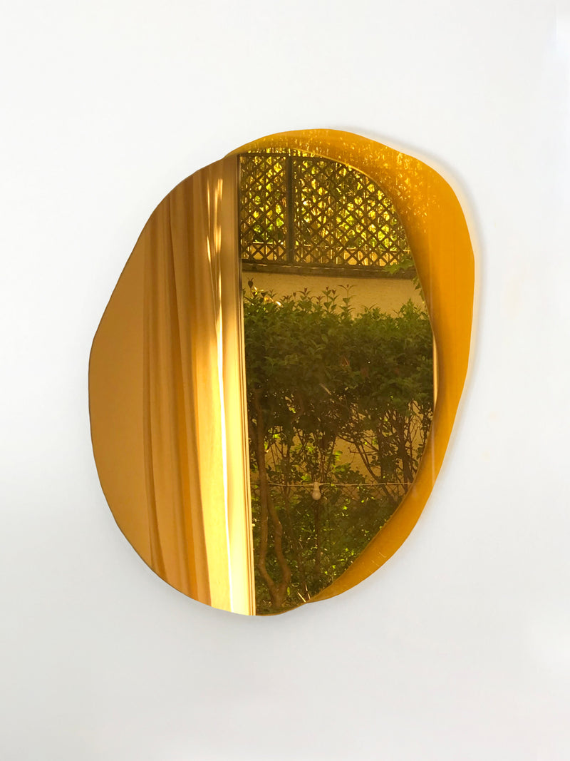 The Mirror Ombrée Gold by Laurène Guarneri, featuring a round, golden frame and a smooth, irregularly shaped edge, hangs on a white wall. The reflection in this handmade poetic mirror shows cream-colored curtains and a view of greenery outside the window, including bushes and a lattice fence.