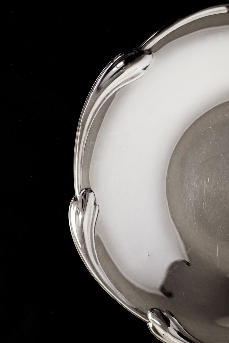 A close-up of the septembre studios Large silver-plated dish with scaled rim, showing its smooth, reflective surface and elegant rim detail against a black background. Only part of this decorative dish is visible.