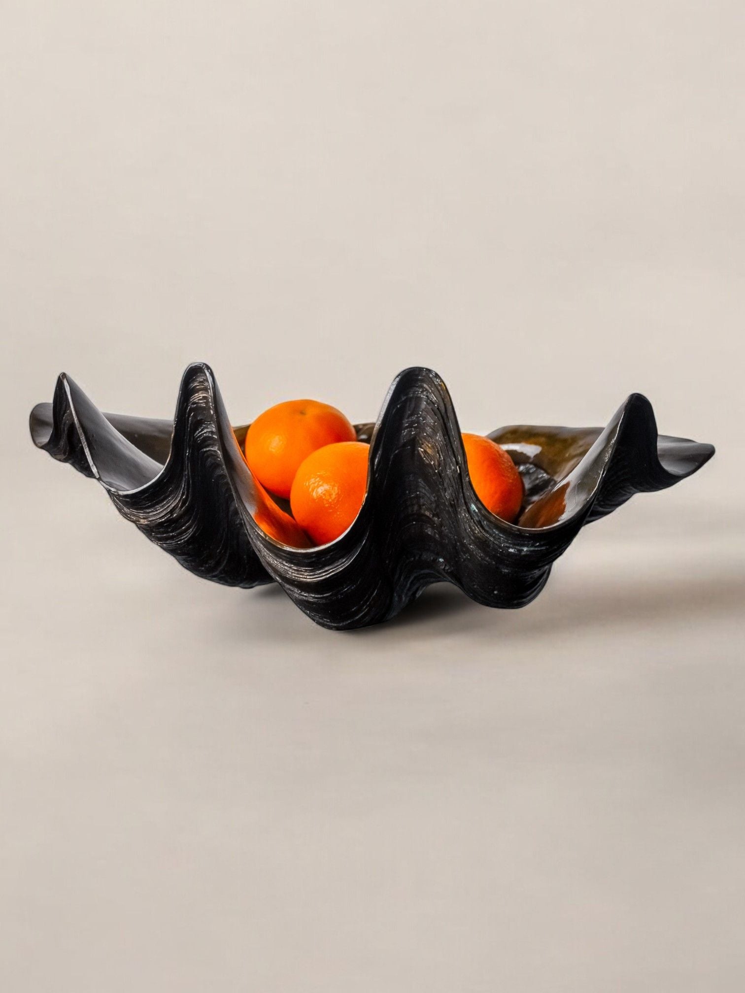 A wavy, black ceramic bowl resembling a scallop shell displays several small oranges. The Large Shell in Plaster from Malamar Studio, with its undulating design, contrasts strikingly with the vibrant fruit inside against the neutral, light beige background.