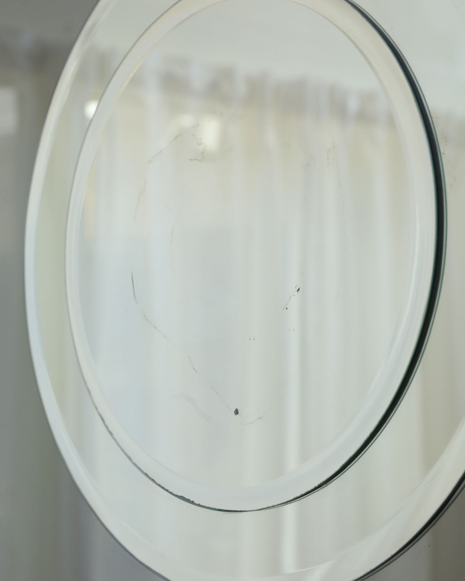 A close-up of Villa’s 1970’s Mirror, a round two-tone smoke mirror featuring visible smudge marks on its surface, set against softly blurred white curtains.