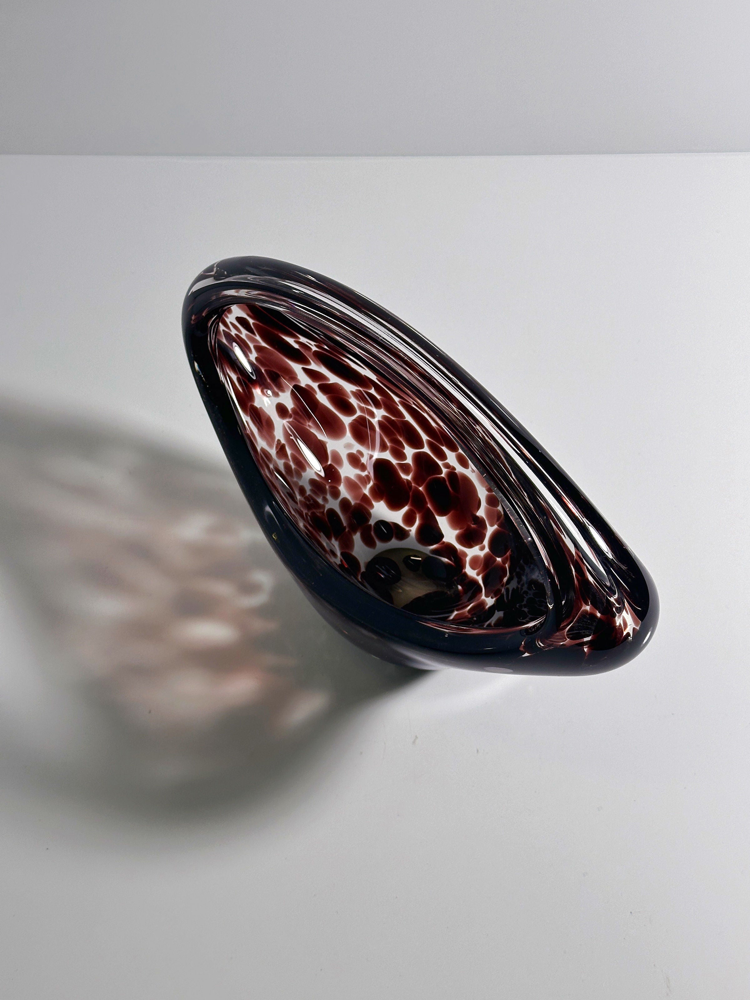 The Dodo Vintage Murano Tortoise Glass Handle Bowl - 1970s features a unique, asymmetrical design with a dark red and white swirling pattern, evoking the classic tortoise appearance. Its glossy texture casts soft shadows on white surfaces, reminiscent of vintage Murano craftsmanship.