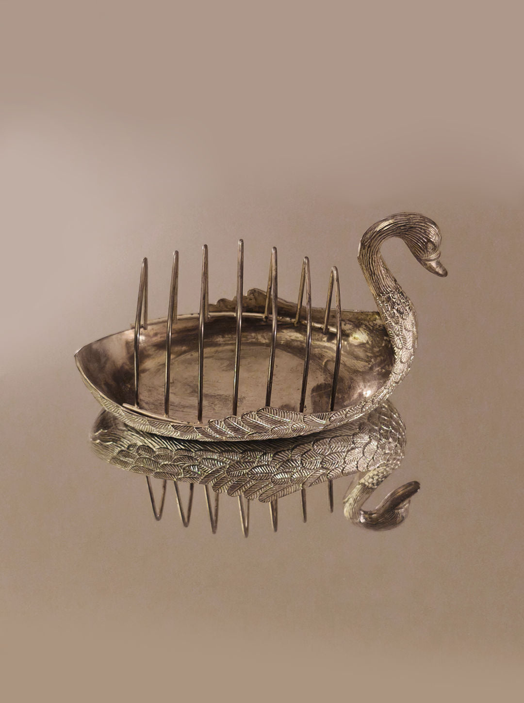 The Swan Toast Rack by Les Objoies is displayed on a reflective surface, creating a mirrored effect. This silver-plated breakfast table accessory is shaped like a swan with intricate detailing, and its elegantly curved neck serves as the handle.