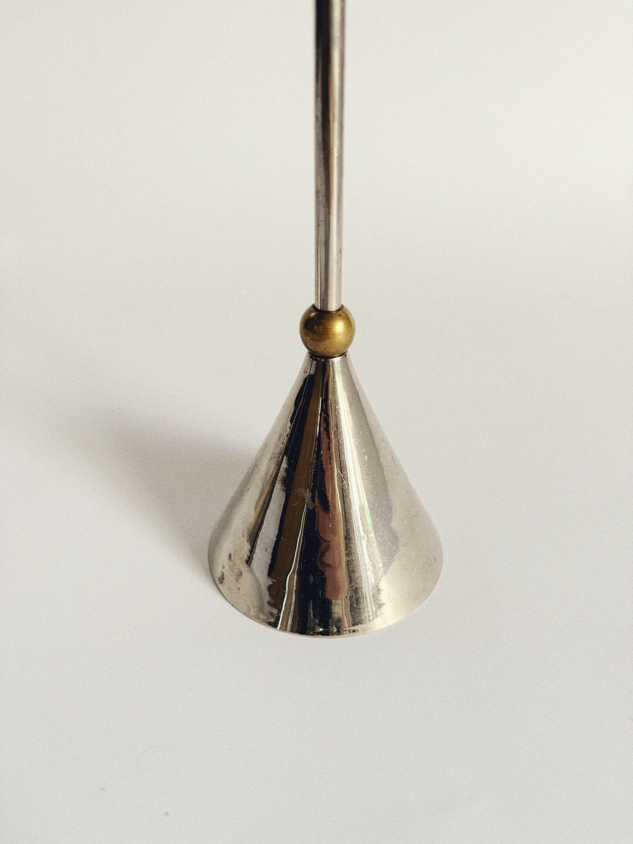 A close-up of one of Huerto's Set of 3 Postmodern Candle Holders, featuring a metallic cone-shaped design with a shiny chrome finish that exudes sleek elegance. A small brass sphere elegantly connects the holder to its vertical rod, set against a plain, light background for visual contrast.