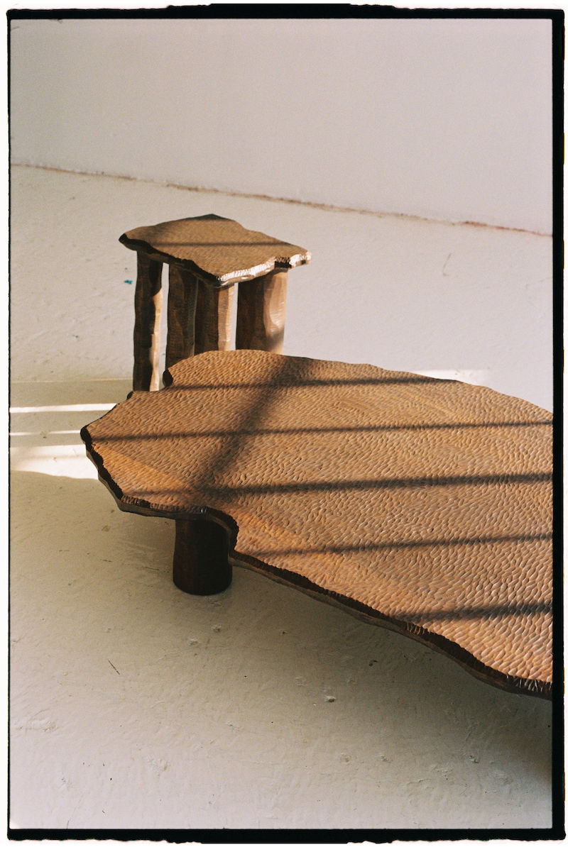 In a minimalistic, sunlit room, an Ombia Studio Mare Side Table and its uniquely shaped wooden coffee table counterpart, crafted from solid white oak with a textured surface, are accented by patterns of shadows from blinds on the light-colored floor.