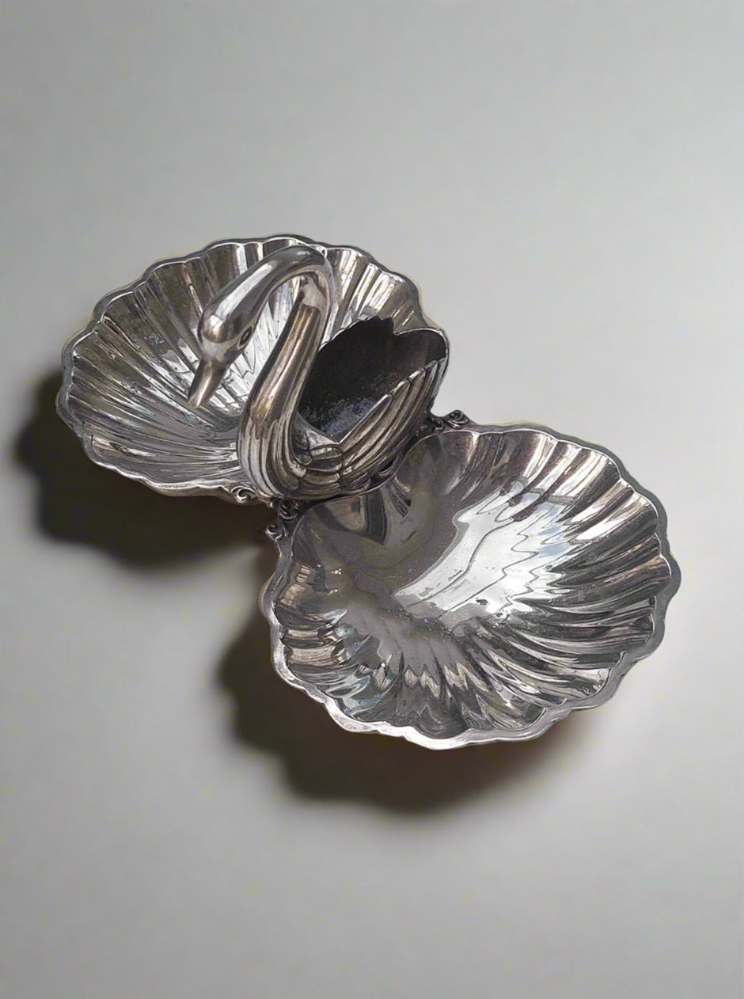 The Les Objoies Shell and Swan Dish is showcased against a white marble surface. This metallic dish, shaped like two shells, features an elegant swan figurine with an arched neck serving as a charming divider between the shells. Its shiny, reflective surface has slight oxidation marks that add character to its gleam.