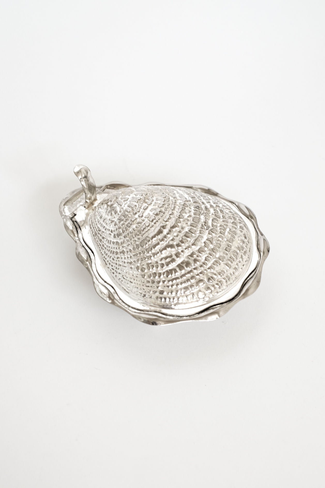 The Shell Butter Dish by septembre studios features a vintage silver oyster shell shape with a textured, ridged surface and a small curved handle, elegantly displayed on a plain white background.