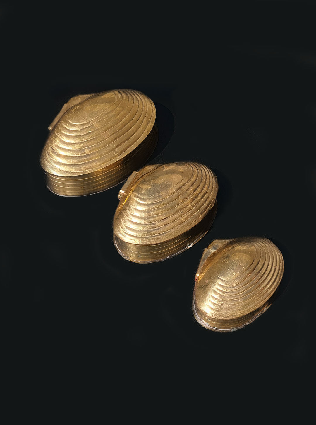 Three polished, gold-colored seashell-shaped objects with a brass gold finish are displayed against a black background, showcasing their ridged textures and shiny surfaces. The Set of 3 Nesting Shell Boxes by Les Objoies are artfully arranged to highlight their exquisite detail and craftsmanship.