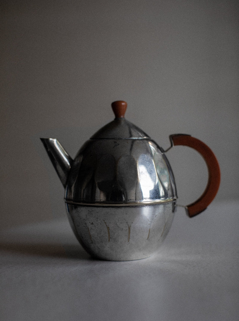 A silver-plated teapot with an orange handle and an orange knob on the lid is displayed on a plain surface. The body of the teapot is rounded with a slightly scalloped design, providing timeless elegance. The background is out of focus, highlighting the Small Teapot by Out For Lunch’s shiny, metallic surface.
