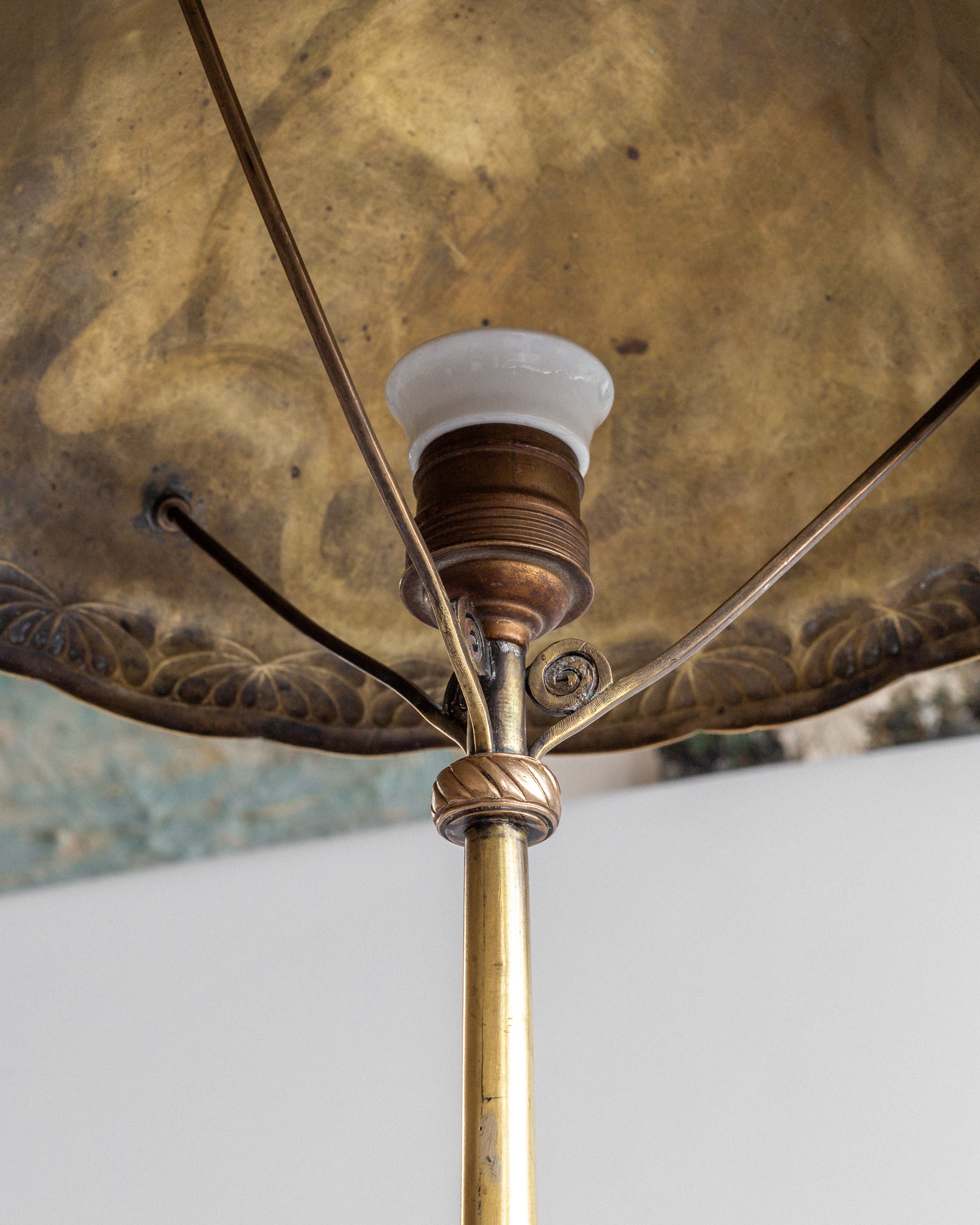 Close-up of an Art Nouveau Brass Table Lamp by Spigel, featuring a vintage brass finish with ornate detailing. Its decorative metal canopy and exposed bulb socket connect through rods to a central stem with floral engravings.