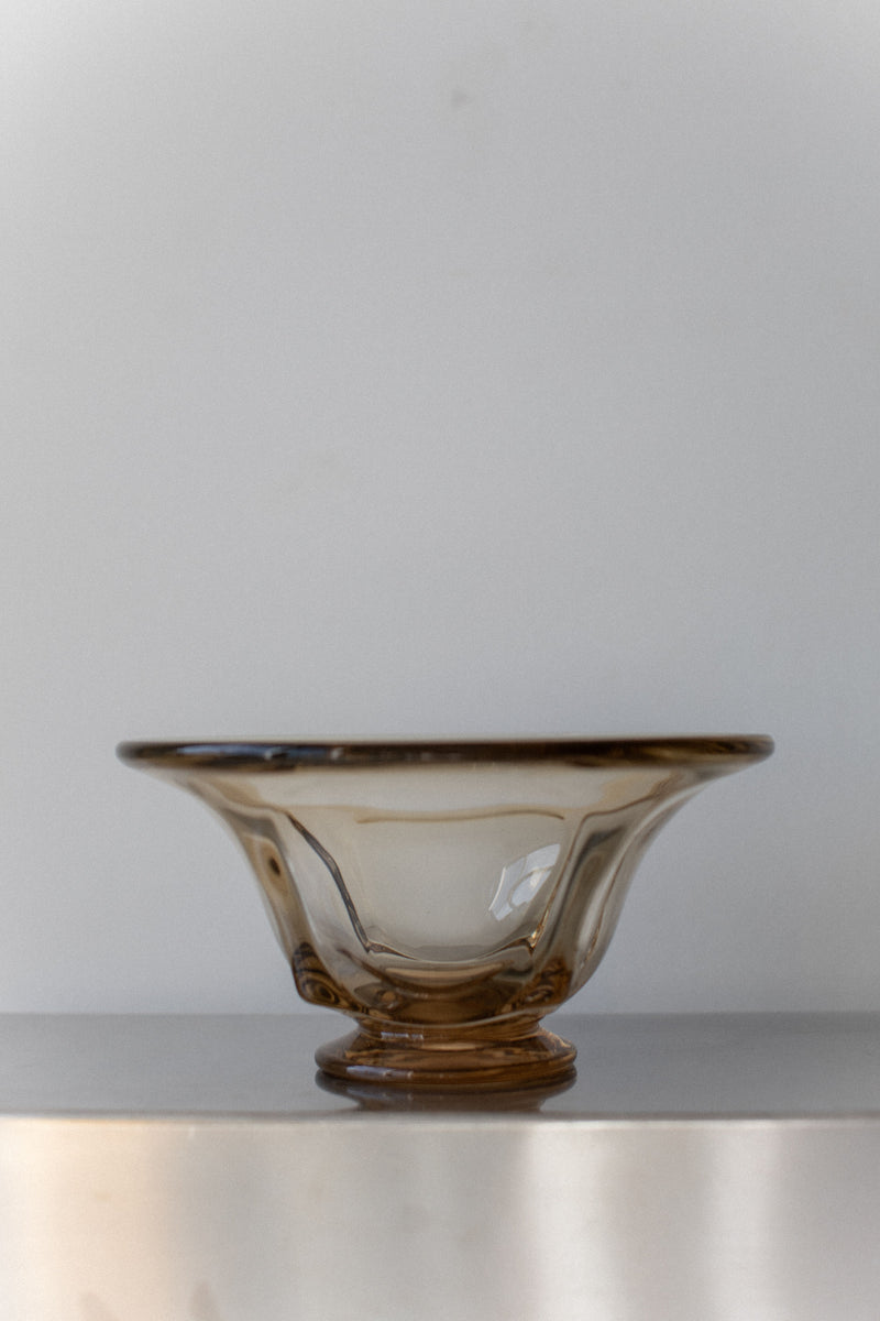 A Vintage Glass Bowl from Out For Lunch, featuring a translucent brown tint and graceful wavy rim, rests on a smooth surface against a light gray background. Embodying mid-century design, its delicate swirling patterns and petite circular base exude timeless charm.