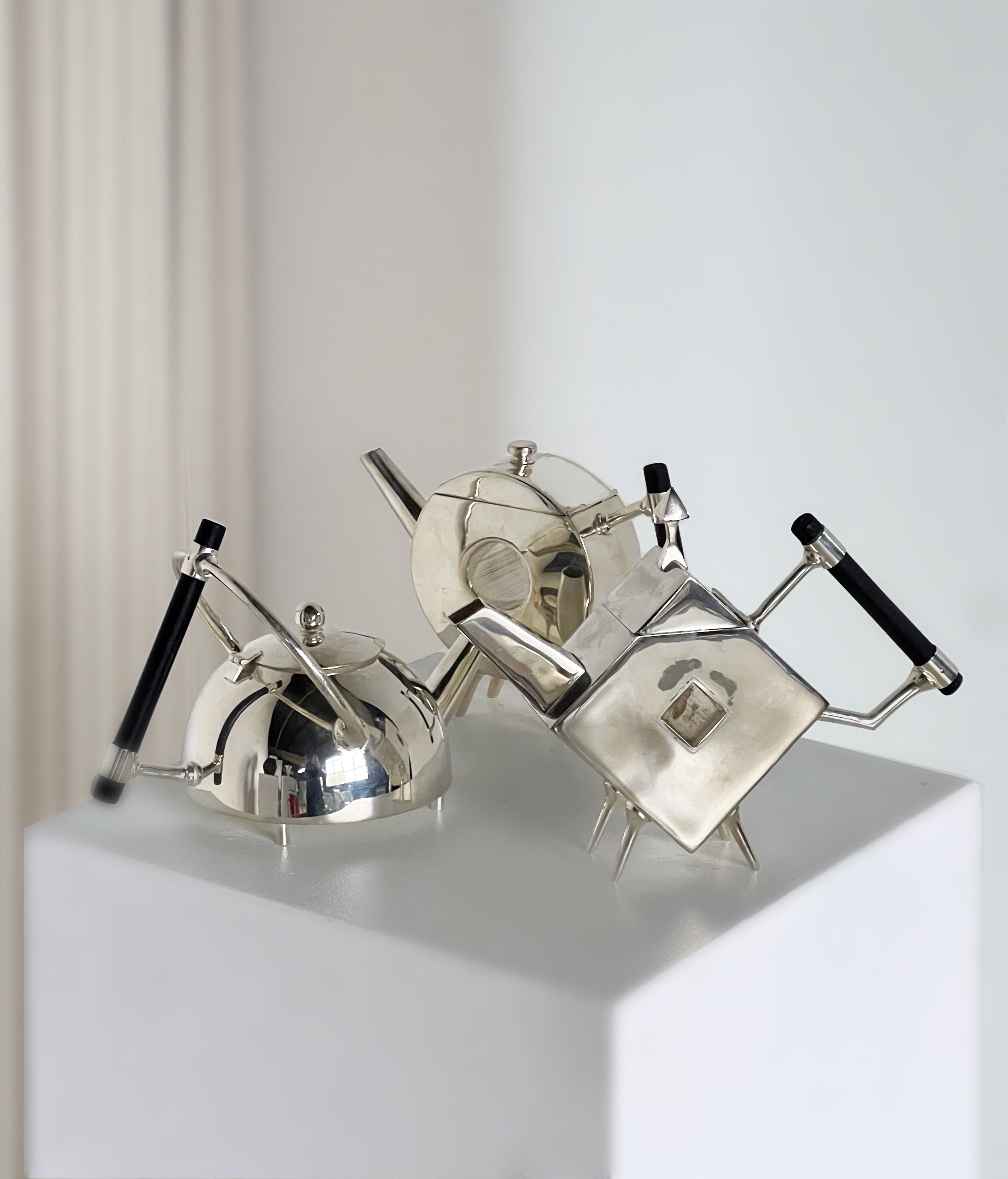 Three modern, silver-plated teapots by Porter Studio, each with a unique geometric shape and black handles, are displayed on a white pedestal. The softly blurred background enhances their design elegance, reminiscent of the Christopher Dresser Ornate Circular Teapot.