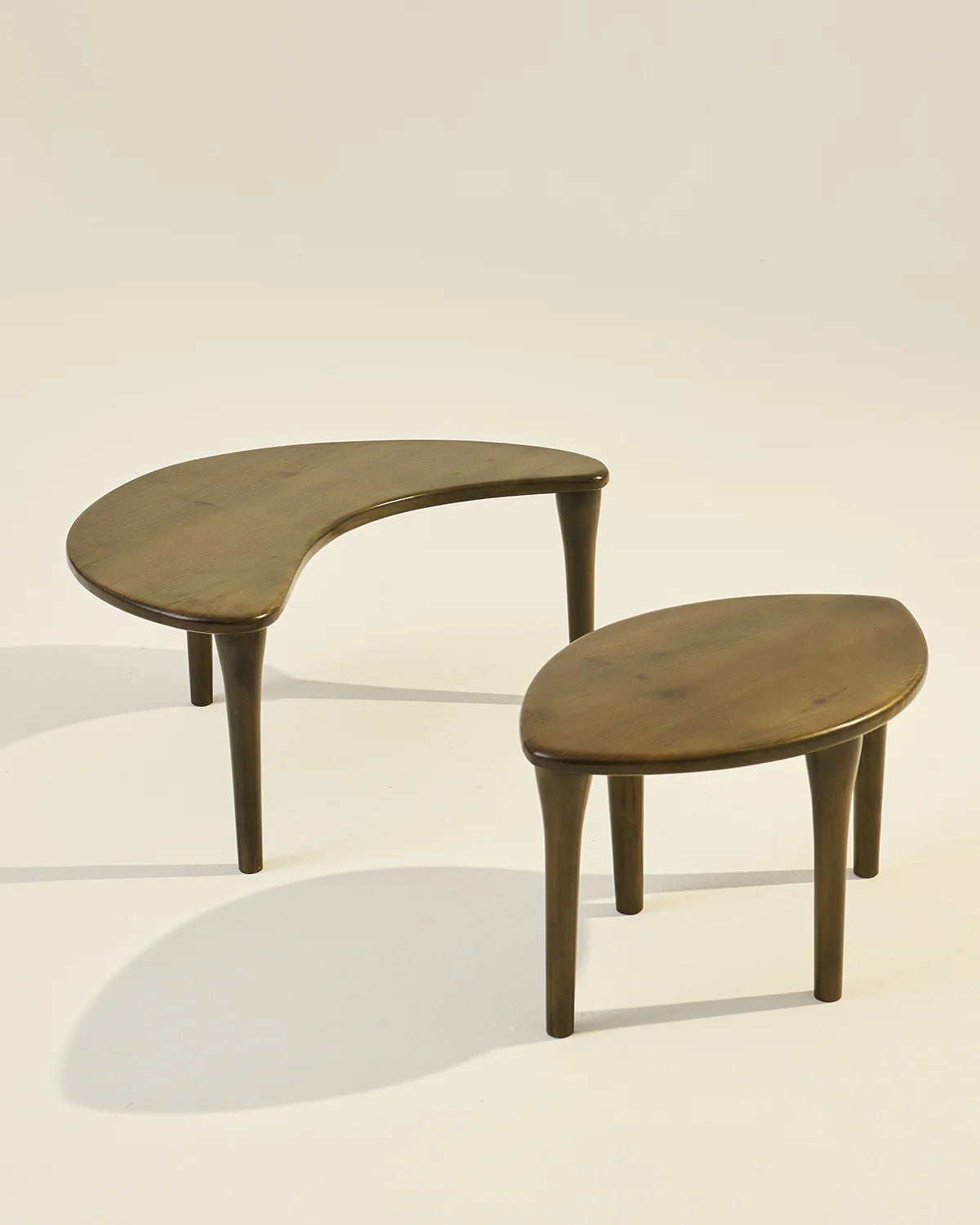 The Crescent Coffee Table Set of 2 by LMNOH features wooden tables with asymmetrical kidney and teardrop shapes crafted from ashwood. Each table has three tapered legs, offering space-saving functionality, set against a neutral background with soft shadows.