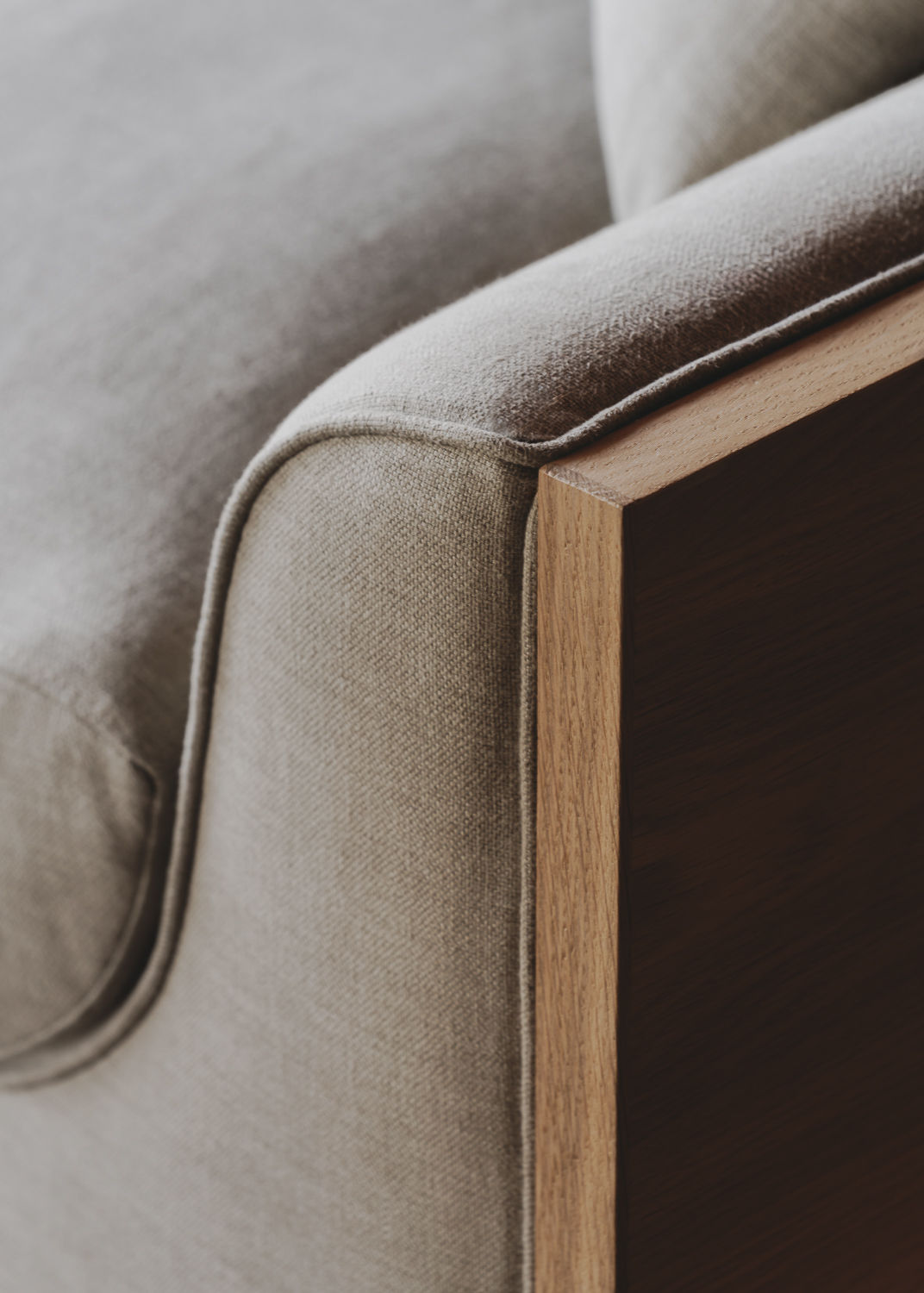 A close-up of the Marina Sofa by ASERIES OF OBSTACLES, SL highlights its light beige fabric and wooden armrest. The textured fabric contrasts with the sleek solid oak frame, showcasing a minimalist design.