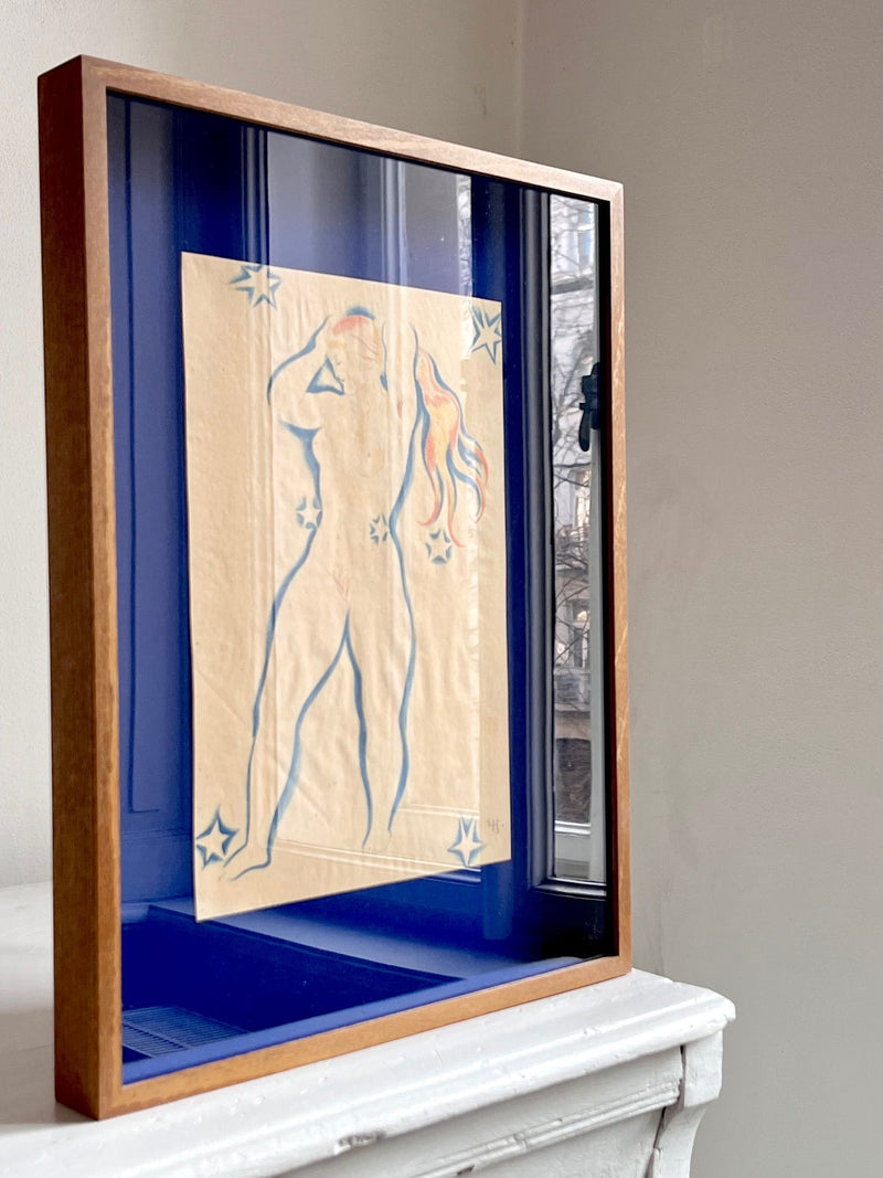 The Médecine 1930s Drawing by Ernst Hansen is a framed original showing a blue-outlined nude with orange and stars on a blue background, displayed on a white mantel beside a window.
