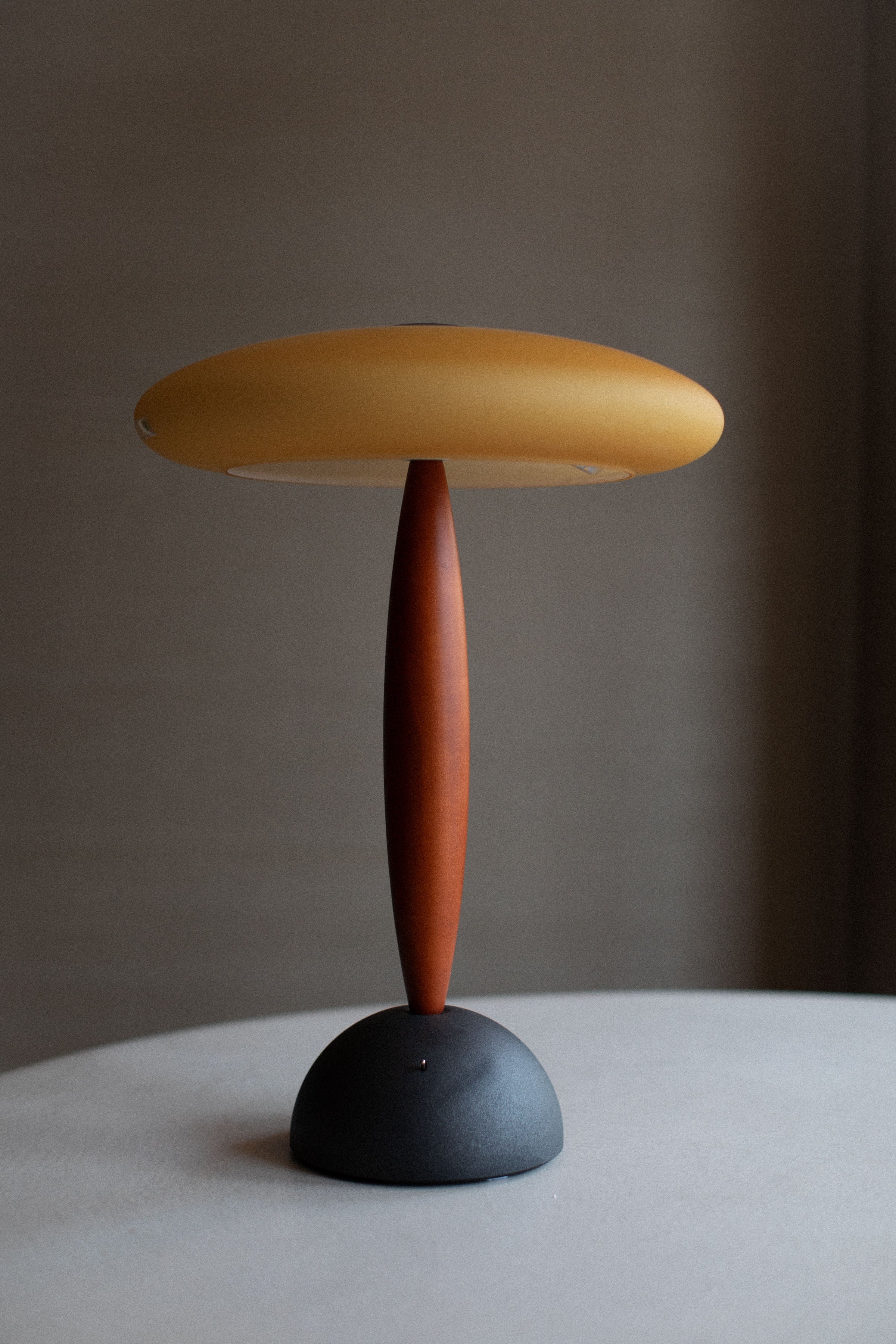 A modern Vintage Murano Lamp with an elliptical yellow shade balanced atop a slender, vertical brown stand, handcrafted by Murano glassmakers, which is mounted on a round, flat black base, against a