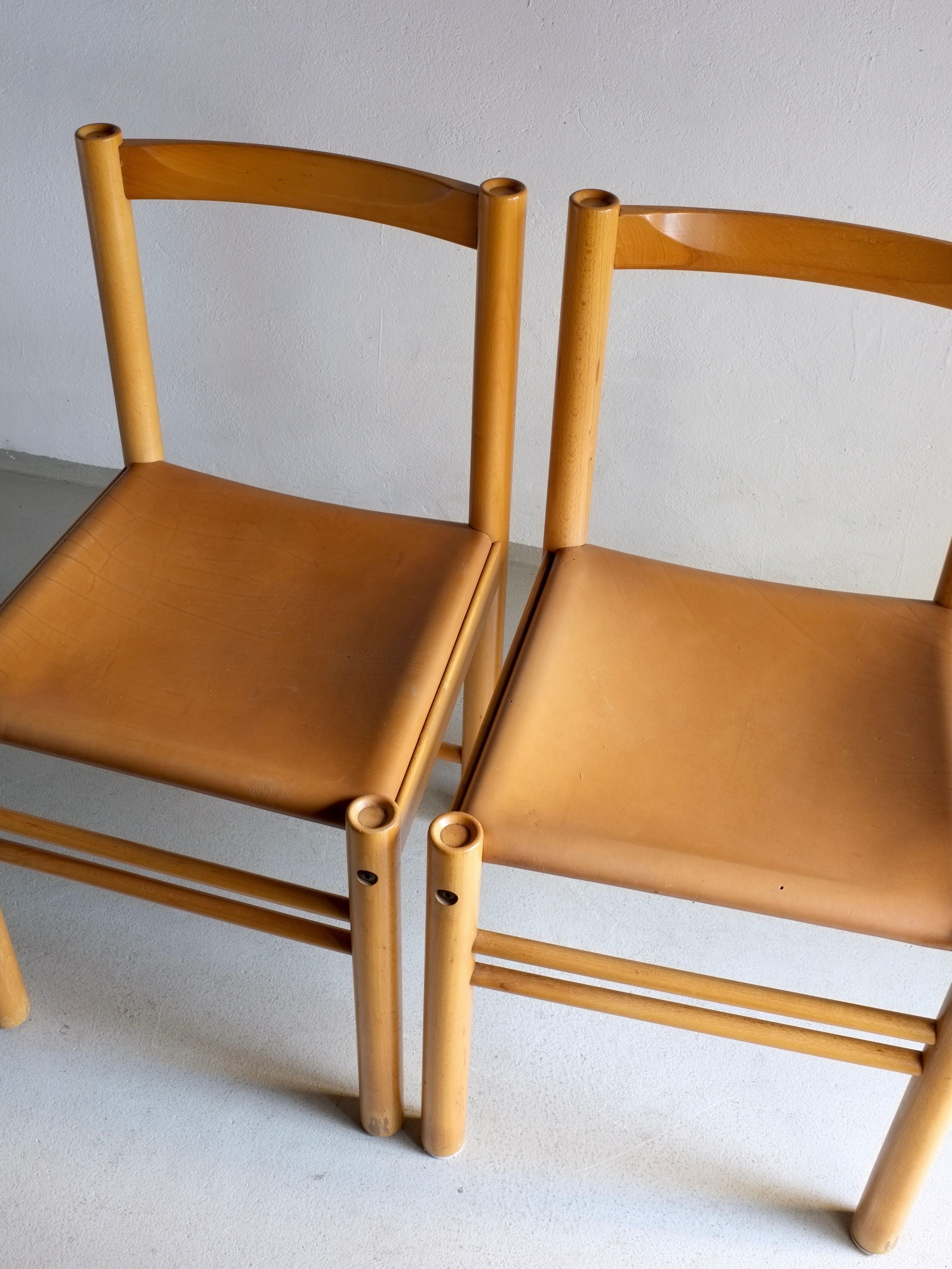 Two chairs from Veter Vintage's collection of 6 Beech Wood Cognac Leather Chairs by Ibisco, crafted in Italy during the 1960s, are placed side by side on a grey floor against a white wall. Their minimalist design features rounded legs and backs, showcasing an elegant vintage aesthetic perfect for any setting.