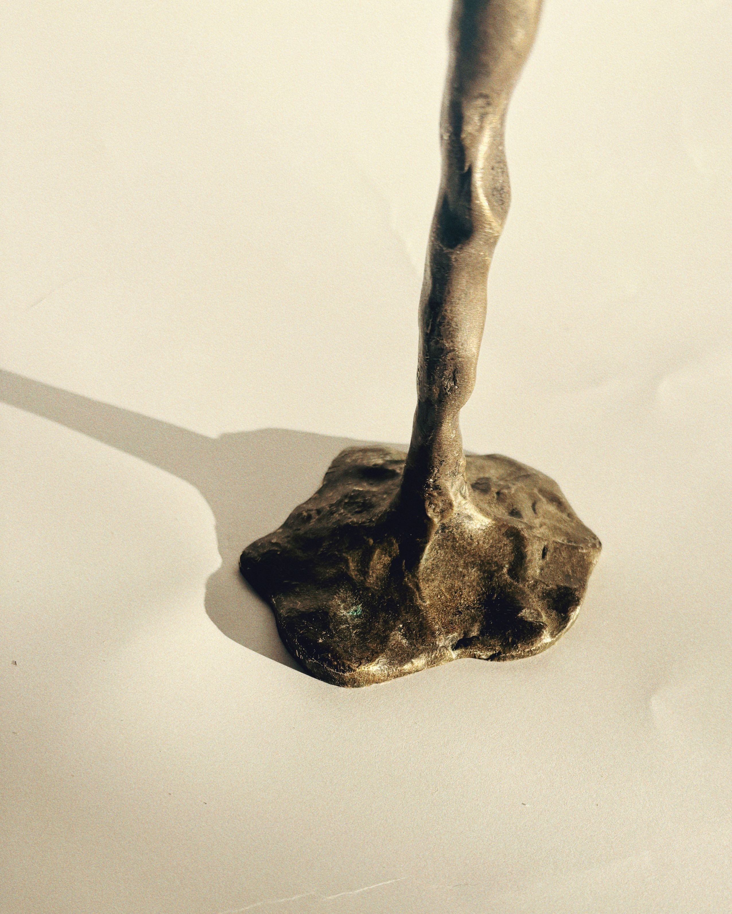 Close-up of a bronze sculpture by Huerto, artist-signed and resembling a twisted rod anchored onto an irregular, textured base. This unique Bronze Candle Holder casts a subtle shadow on the light-colored surface beneath it, showcasing its avant-garde design.