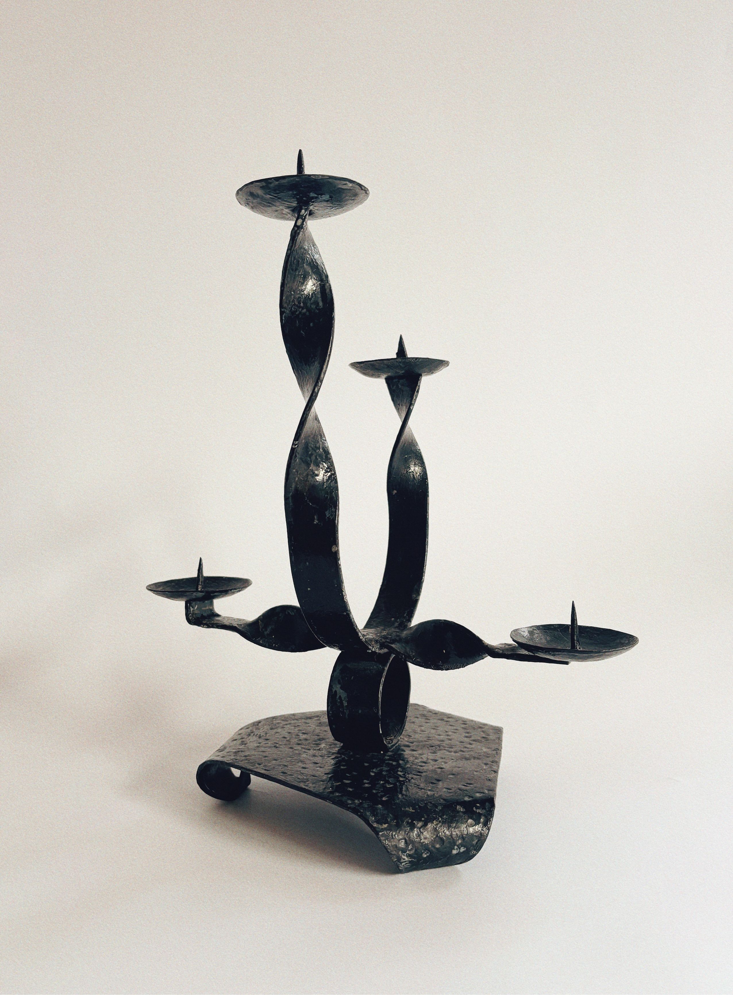 The Huerto Black Metal Candlestick, featuring twisted ribbon-style arms that elegantly extend outward with plates for holding candles, is set against a plain white background. The base of this handcrafted candlestick is flat with a gracefully curved edge.