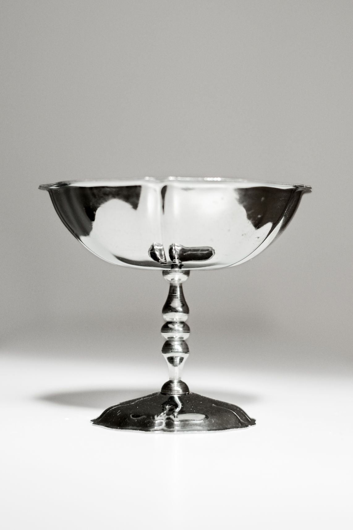 A set of six shiny stainless steel ice coupes by septembre studios, each featuring a wide bowl and decorative stem, stands on a white surface with vintage appeal against a plain gray background.