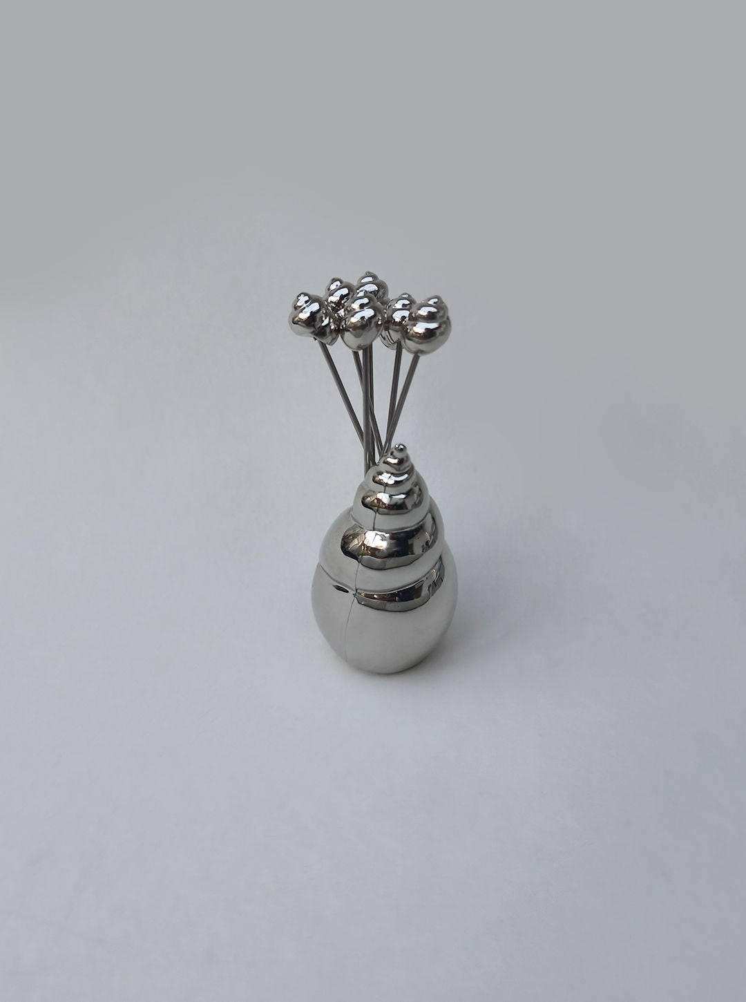 The Les Objoies Shell Cocktails Picks set features a shiny silver spiral shell holder with several ocean-inspired metallic picks, each topped with a small rounded ornament, displayed against a light gray background.