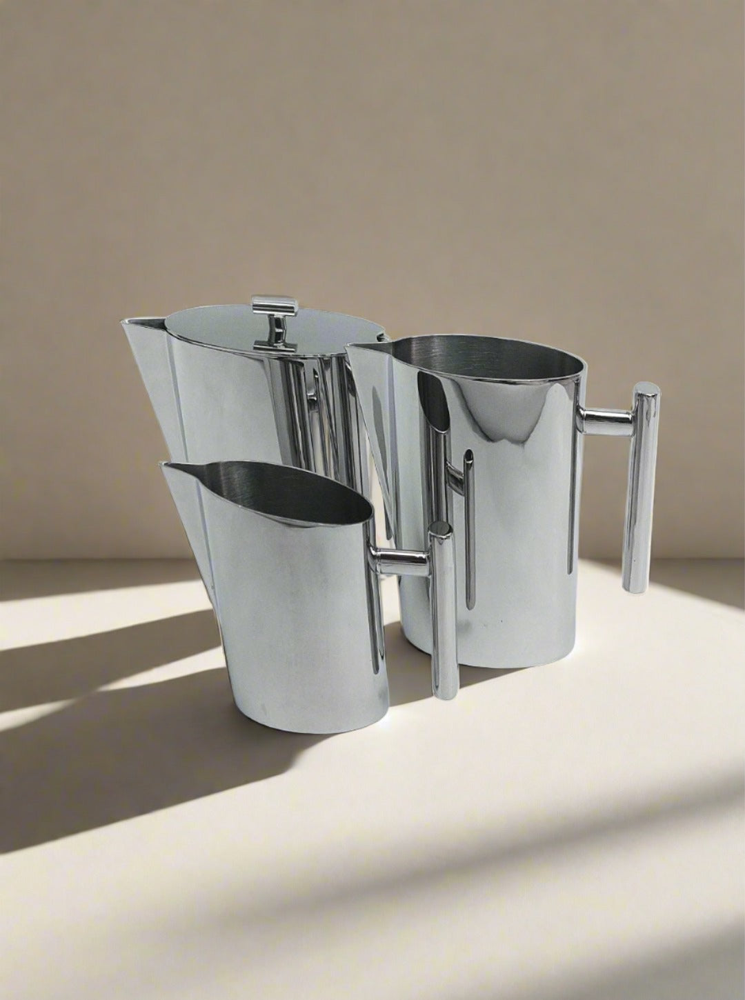 A set of three Les Objoies Vintage Coffee Set polished stainless steel pitchers, resembling a sleek coffee or tea set, are arranged on a light-colored surface. The pitchers have modern designs with long cylindrical handles. Sunlight casts soft shadows from the pitchers onto the surface.