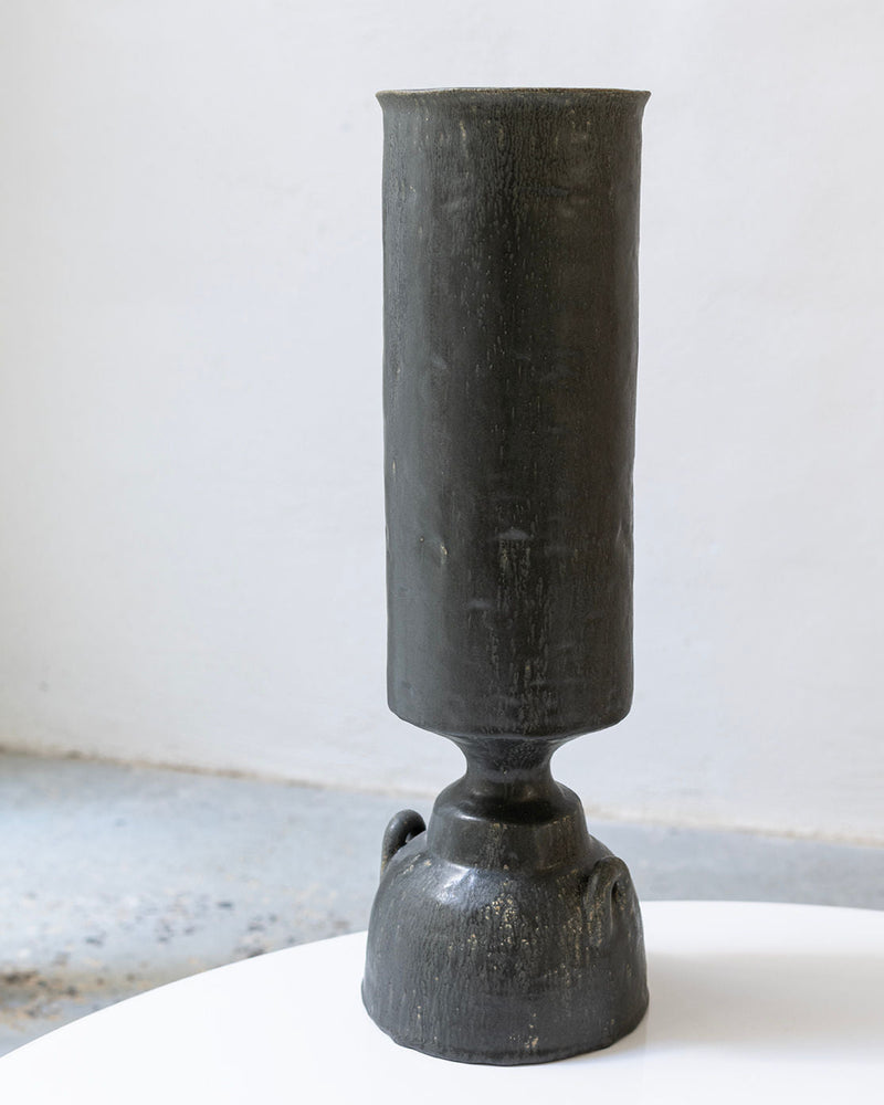The Isolated n.32 Vase by Canoa Lab is a tall, dark ceramic piece with a cylindrical top and rounded, textured base, displayed on a white surface against a plain light background.