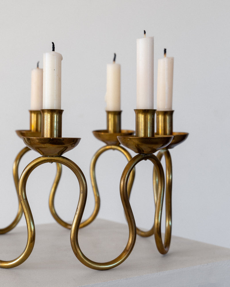 A 1940s Scandinavian Brass Candleholder by Spigel, featuring an elegant loop design reminiscent of vintage styles, holds six tall white candles with blackened wicks on a light surface, reflecting prior use and echoing Lars Holmström's Scandinavian influences.