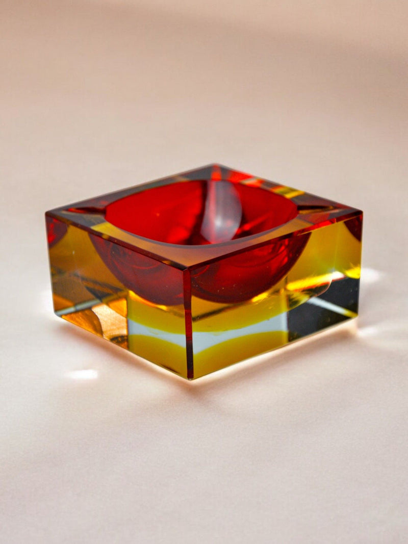 The Sommerso Ashtray by Seguso, Murano, 70s from Malamar Studio is a transparent rectangular block encasing a red sphere. Amber and yellow hues reflect off its sides, creating a vibrant effect reminiscent of the sommerso technique when placed on a light surface.