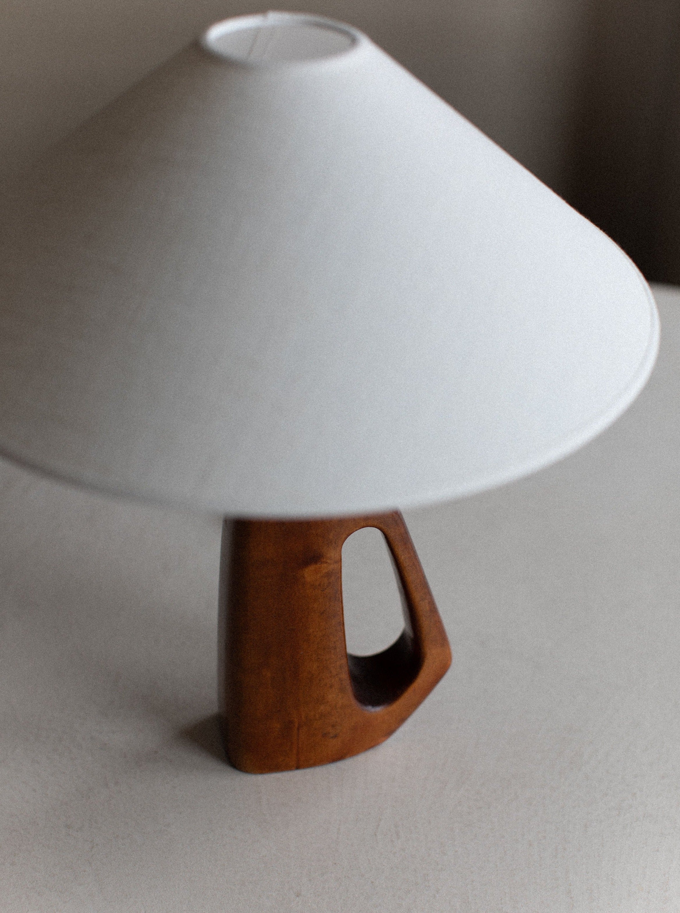 A **1960's Wooden Lamp** with a white conical shade and a unique wooden base featuring hand-carved details and a triangular cutout sits on a light-colored surface. The lamp's mid-century design is modern and minimalist, creating a stylish decor piece by **Out For Lunch**.