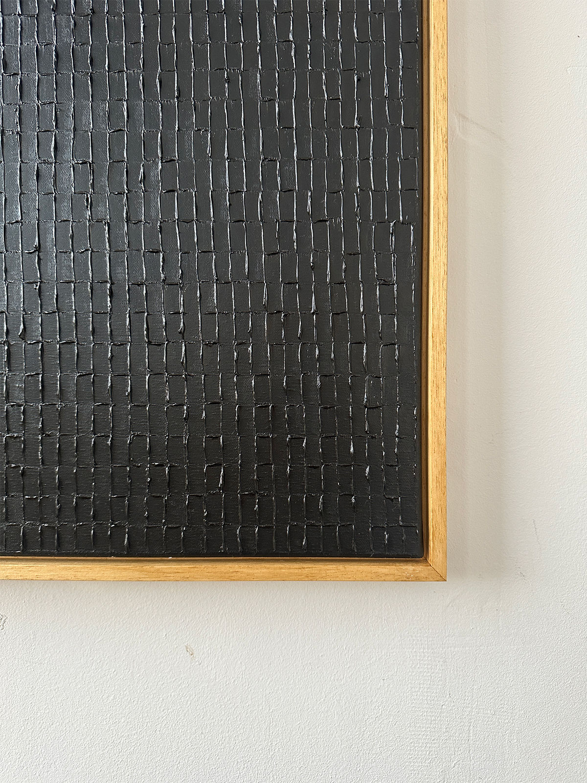 A close-up of "Lamp Black," an oil on canvas by Roi Elmaliah, features a textured black surface with a grid pattern. It's framed in light wood reminiscent of Amsterdam's minimalist style and positioned against a plain, off-white wall, evoking a modern Dutch aesthetic.