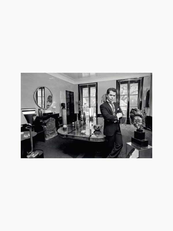 A man in a suit leans against a large wooden desk in a well-furnished, elegant office. The room, reminiscent of Karl Lagerfeld: A Life in Houses by Maison Plage, features modern decor with sculptures, vases, large windows with curtains, and a round mirror on the wall. The overall atmosphere is sophisticated and professional.