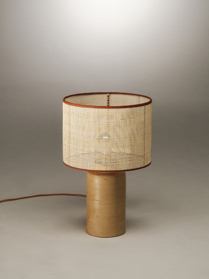 A modern table lamp with a cylindrical wooden base and a wide, cylindrical lampshade made of woven wicker material, reminiscent of classic rattan lamps, highlighted by a thin, brown border. The Sonora Rattan Table Lamp by Nassi Lamps sits on a plain, light-colored surface, with its power cord extending to the left.
