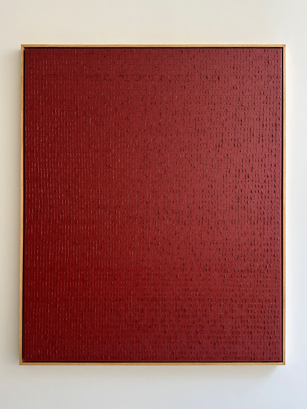 A square art piece featuring a solid red background with vertical textured lines, framed in a simple light wood frame, hanging on a neutral-colored wall. Inspired by the Dansaekhwa movement, the texture adds depth and variation to the otherwise uniform surface. The piece is titled "Indian Red - Oil on canvas" by Roi Elmaliah.