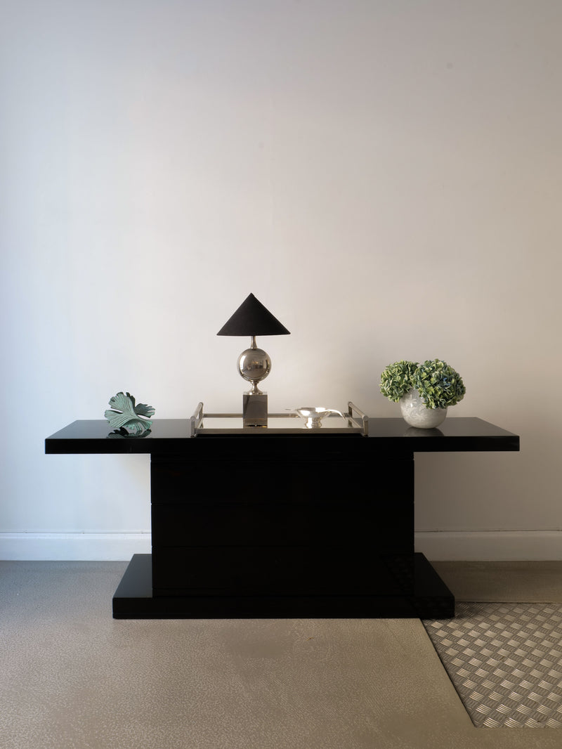 A minimalist setup includes a sleek black table with a geometric lamp, Collection Apart's Bronze Ginko Biloba Sculpture, a small bowl of trinkets, and green hydrangeas in a vase against a plain light wall.