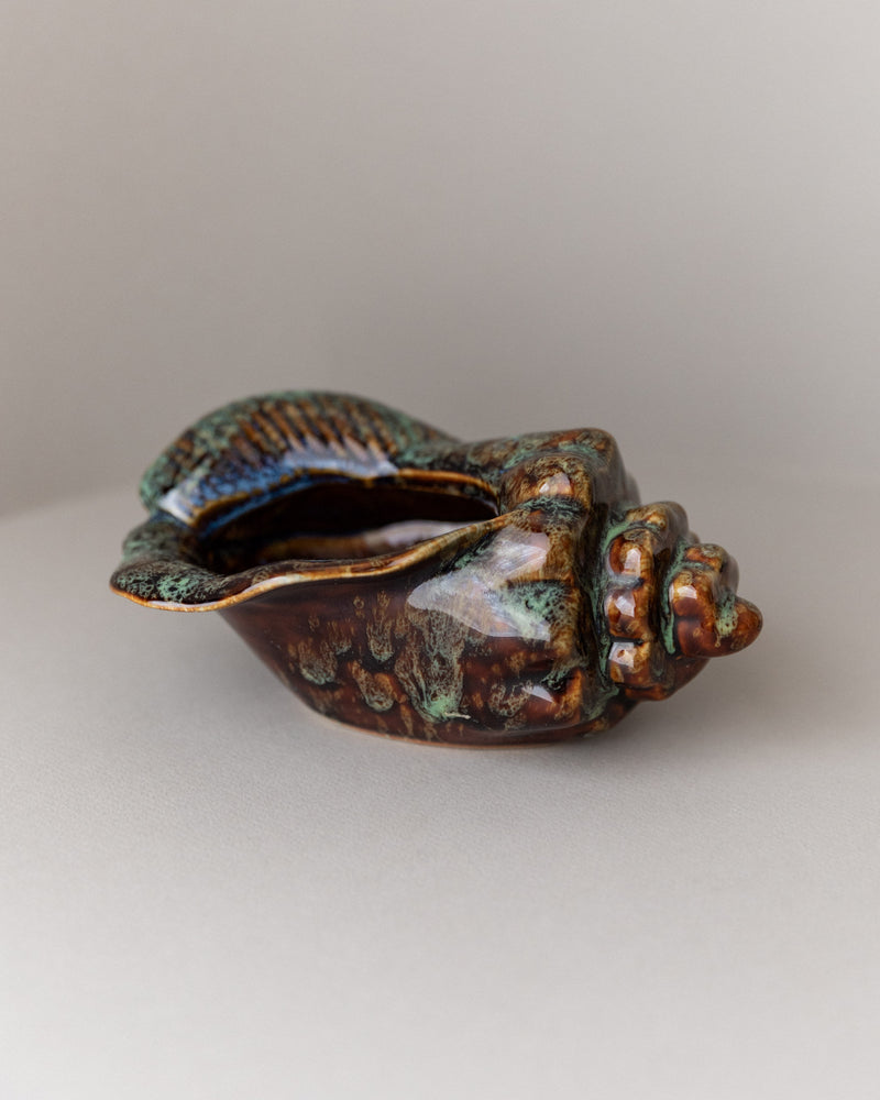 The Bottega Jacobs Ceramic Bowl Shell Shape from Studio Keramik Italy 70s, with a glossy brown and green glaze, intricate textures, and a captivating spiral pattern, elegantly sits on a light gray surface.