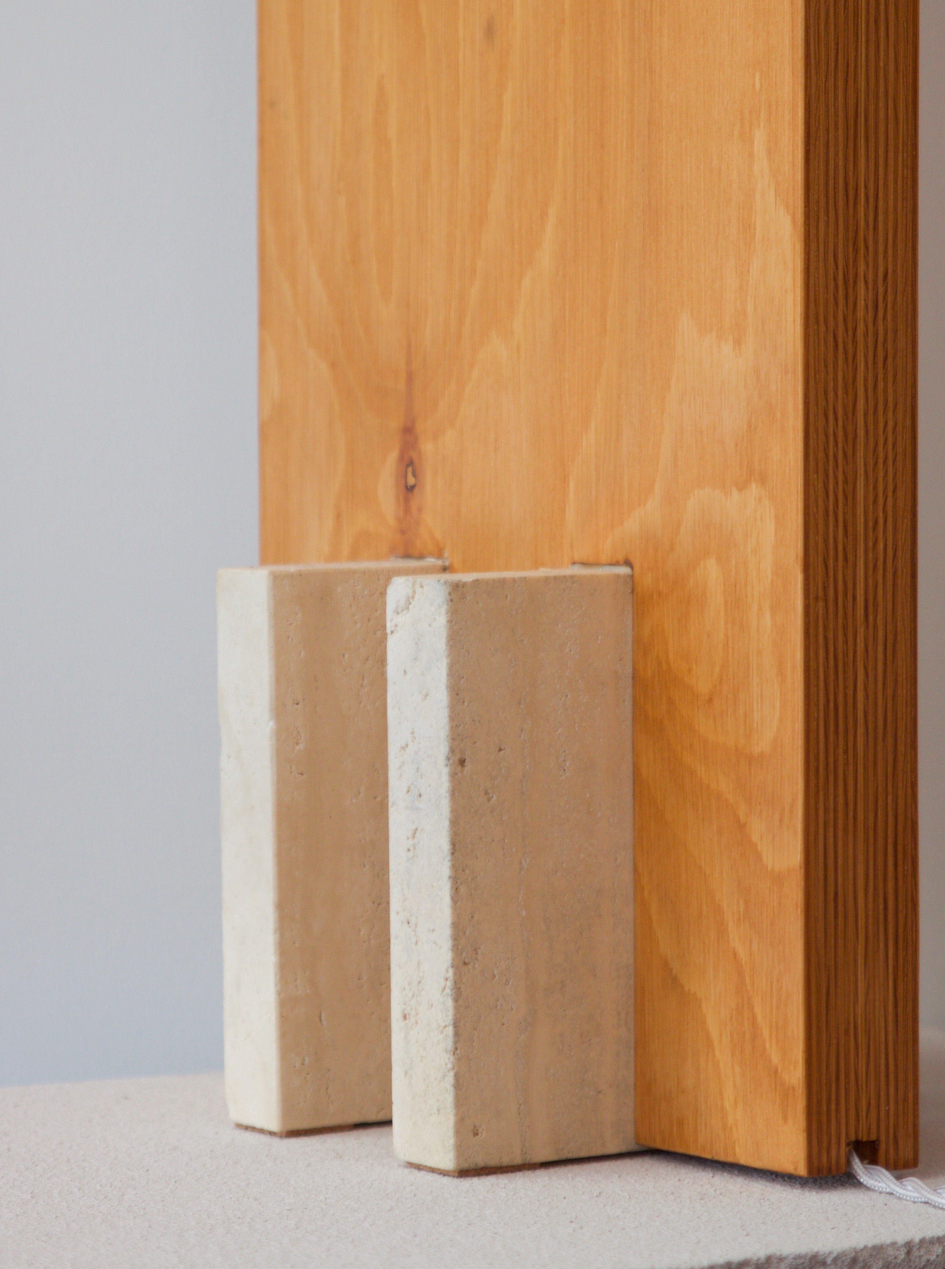 Two Galileo 2.3 beige bricks support a vertical wooden plank against a plain light-colored wall and floor lamp.