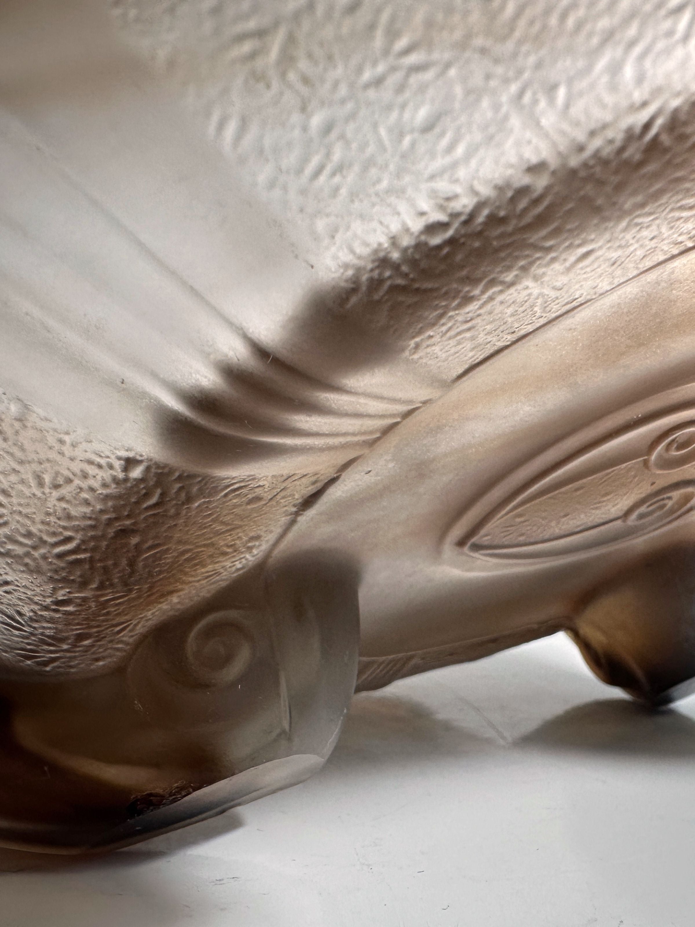 Close-up of the textured surface of a German Art Deco Cloud Glass Bowl circa 1930 by Dodo Vintage, featuring swirling patterns and curves in a light brown hue, elegantly resting on a smooth white surface.