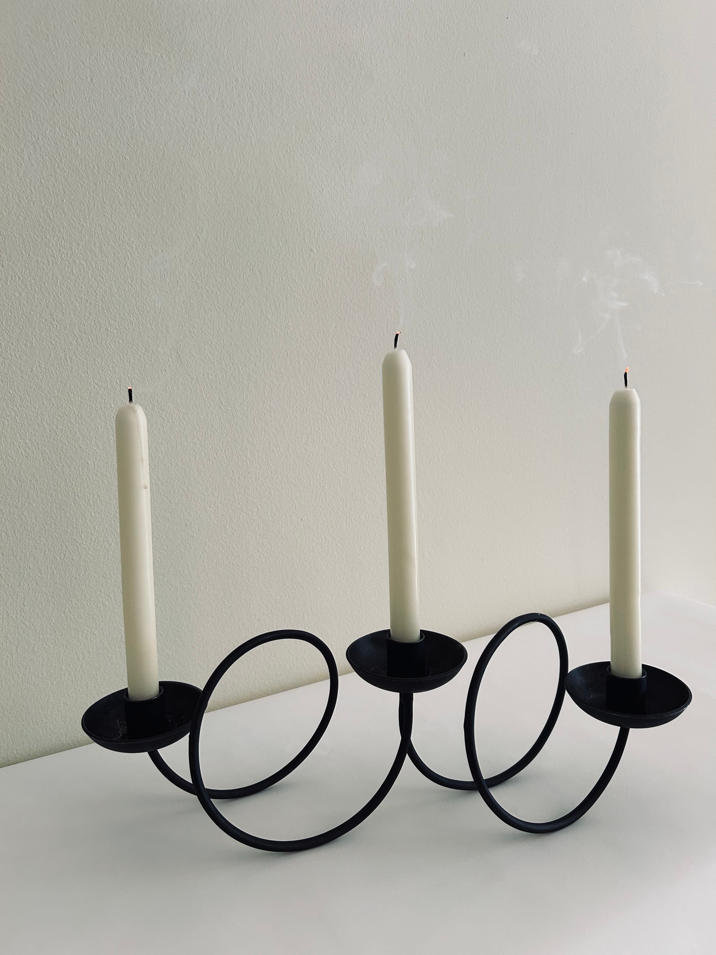 The Brutalist Swirl Candle Holder by Flock Studio features a contemporary metal design with geometric loops, elegantly holding three white candles. Positioned on a white surface with a light backdrop, two of the candles are alight, their delicate flames dancing, while faint trails of smoke drift upward from the recently extinguished third wick.