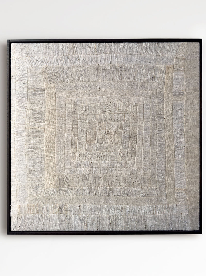The 'To Hold', 2025 by Bec Kirby, is a textured beige textile art piece with a log cabin patchwork style. It features an intricate concentric square pattern in subtle tonal variations, framed in sleek black for a rustic and natural appearance.