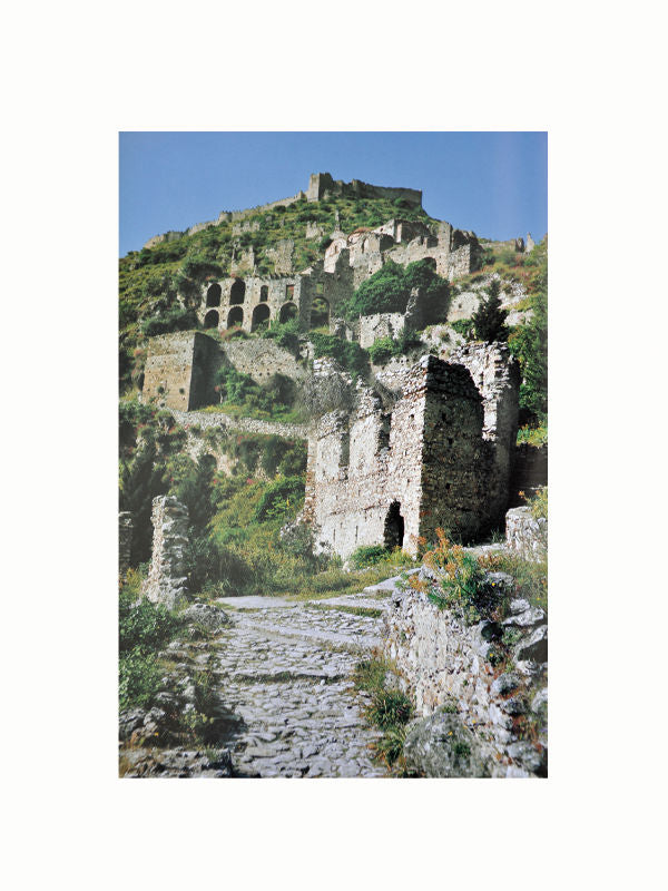 The image shows an ancient, ruined stone fortress on a hill, echoing the grandeur of Greek history. The structures, partially crumbled, are surrounded by greenery. An old cobblestone path winds through the ruins, leading up the hill under a clear blue sky, captured beautifully in "The Greek World: Classical, Byzantine, and Modern" by Maison Plage.