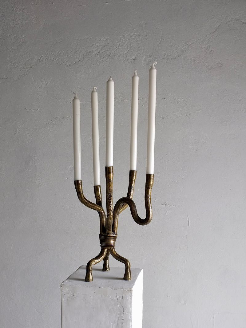 A Brutalist Brass Candelabra from the 1970s by Veter Vintage, featuring five slender, unlit white candles on a brass structure with curved and intertwined arms, stands on a white pedestal against a plain, textured grey background. This elegant piece, hailing from the Netherlands, showcases an intricate design in vintage condition.