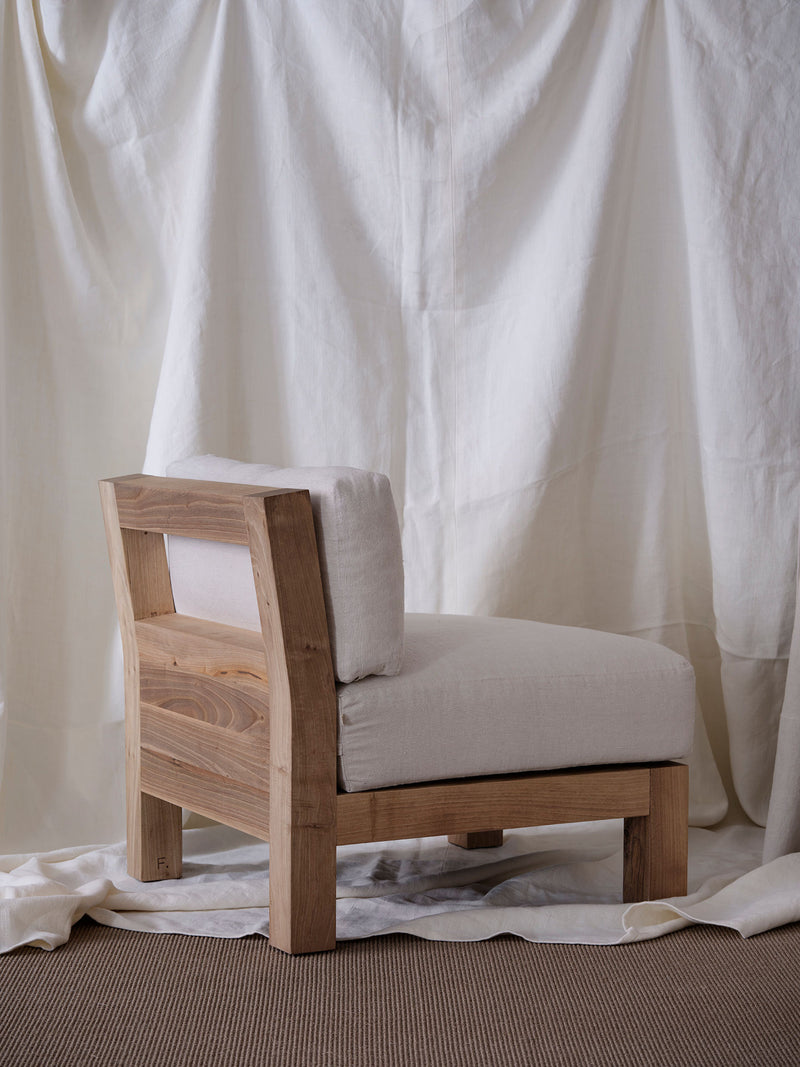 The "Forma" Armchair by FEBRERO SHOP, with its solid wood frame and light beige cushions, rests on a brown carpet against an off-white fabric. Its minimalist design features square lines and a low seating profile, blending comfort and style seamlessly.