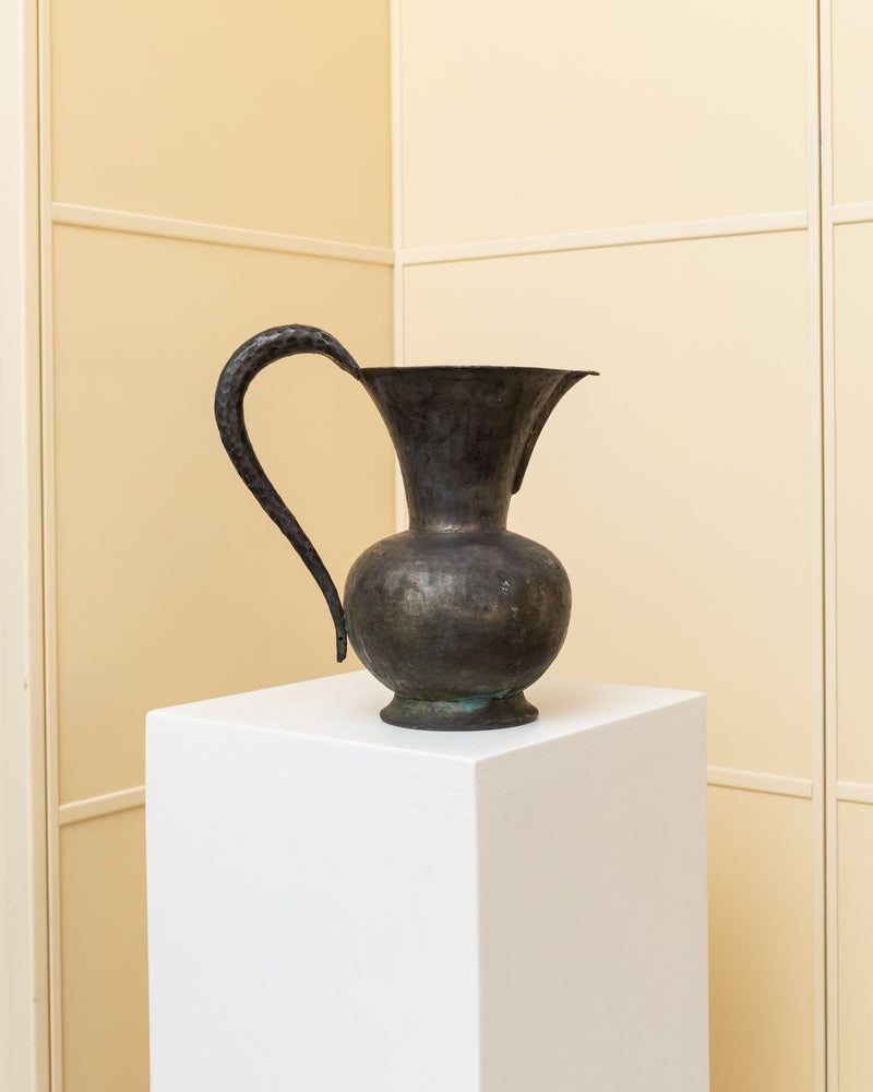 The Vintage metal Carafe - Rameria Lando Rinaldini 1950s from Bottega Jacobs is elegantly displayed on a white pedestal against a cream-colored wall. Its curved handle and surface flaunt an aged patina, enhancing the antique aesthetic with handcrafted details typical of its Italian craftsmanship.