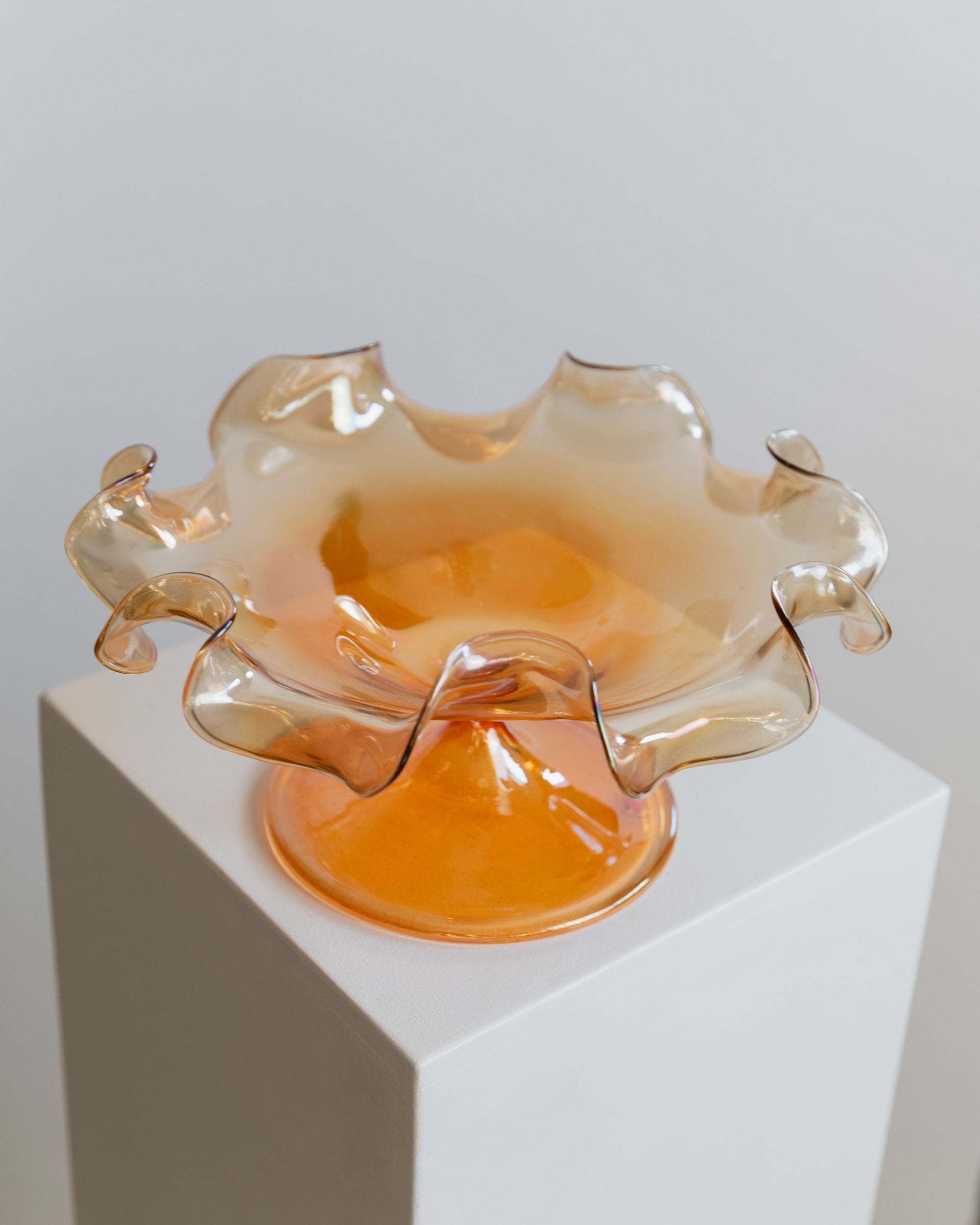 Displayed on a white pedestal against a plain background, the Murano Glass Bowl by Parise Vetro 70s from Bottega Jacobs is a vintage, translucent piece featuring peach hues and wavy edges. Its intricate scalloped design exhibits a subtle orange iridescence, making it an elegant and artistic decorative item.