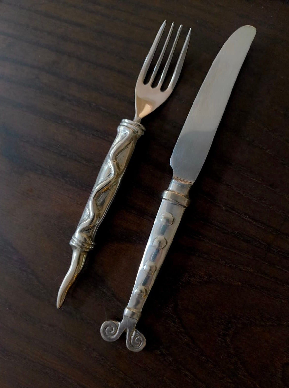 A silver fork and knife set from the Médecine's Rare Vintage Cutlery collection with ornate handles is placed on a dark wooden surface. The fork boasts a twisted handle design, while the knife showcases a geometric pattern complemented by a scroll design at the base of its handle. This elegant set includes 4 forks and 4 knives, featuring durable stainless steel blades.